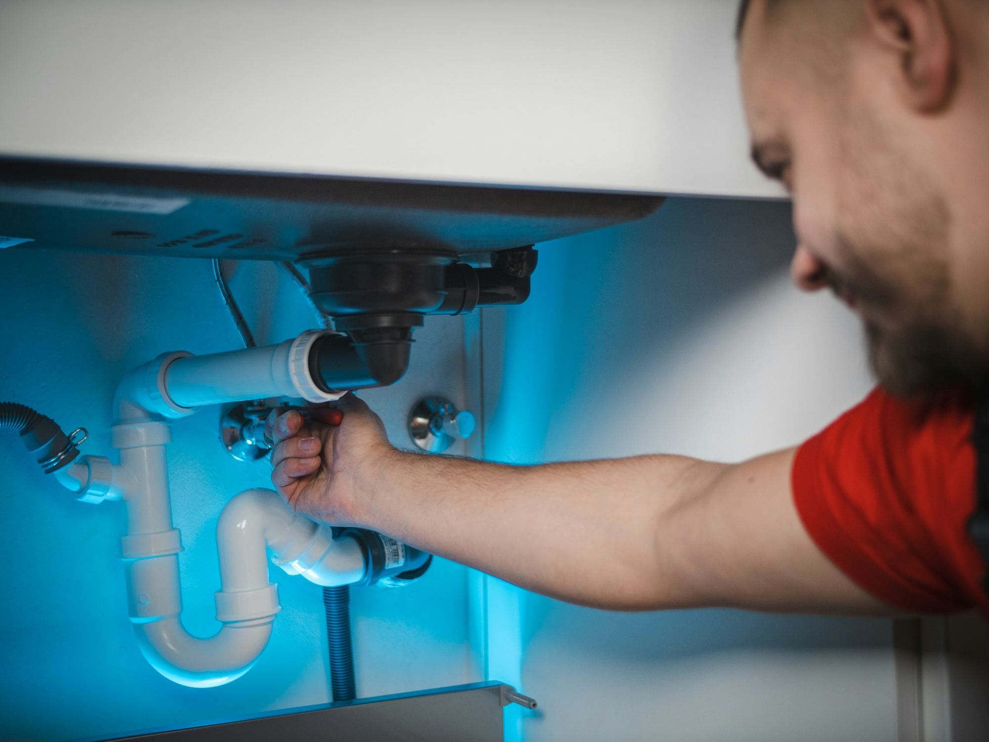A plumber in a red shirt tightening a white plastic waste pipe under blue light